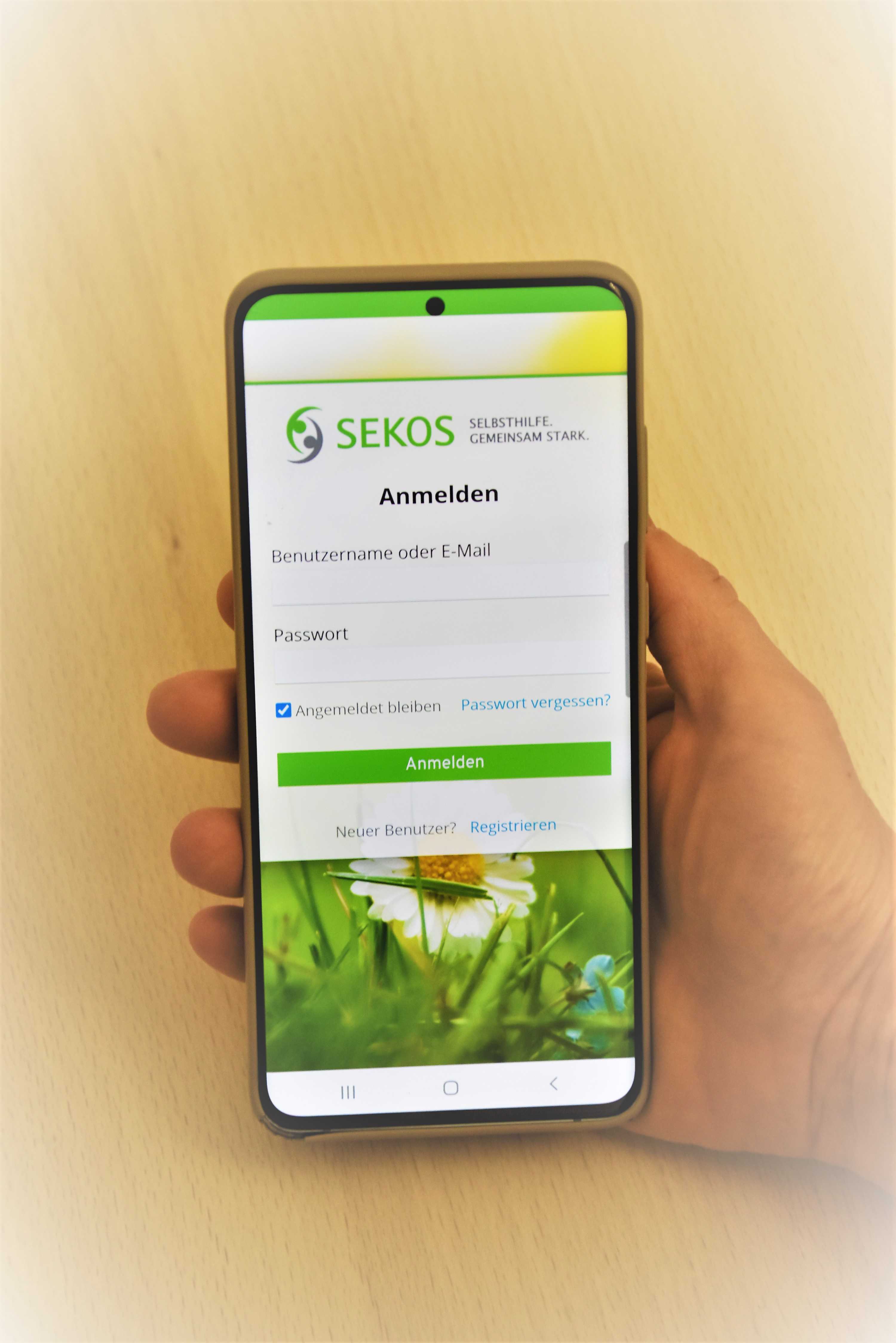 Ein Smartphone in einer Hand. Auf dem Smartphone-Bildschirm ist die Anmeldeseite der App von SEKOS Gelnhausen zu sehen.